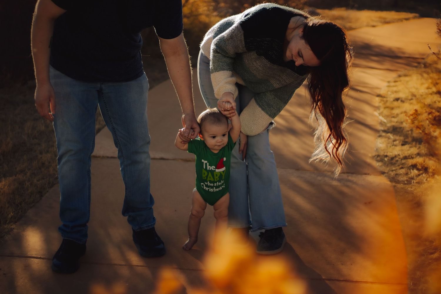 Parents helping toddler take first steps on a sunlit path during golden hour fall family photo session.