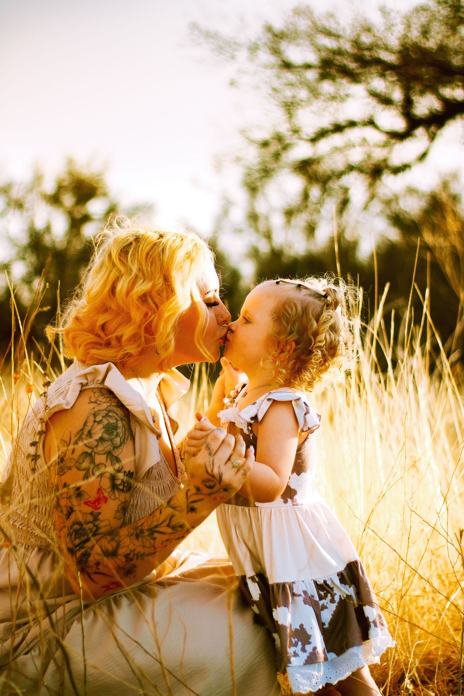 Tattooed blonde mother sharing a kiss with curly-haired toddler daughter in sunny golden field.