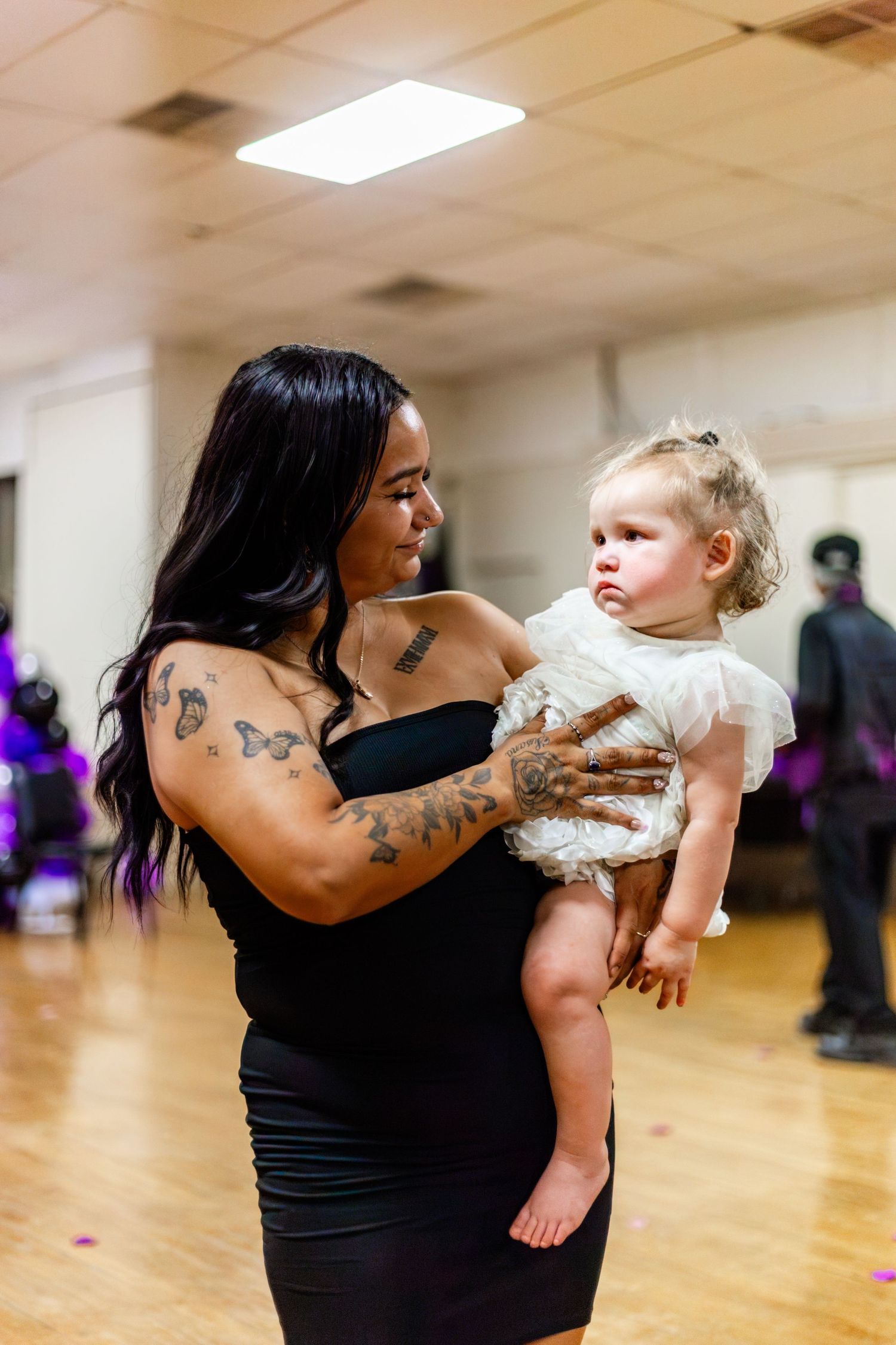 Tattooed woman in black dress holding a baby girl in white outfit at an indoor event with purple decorations.