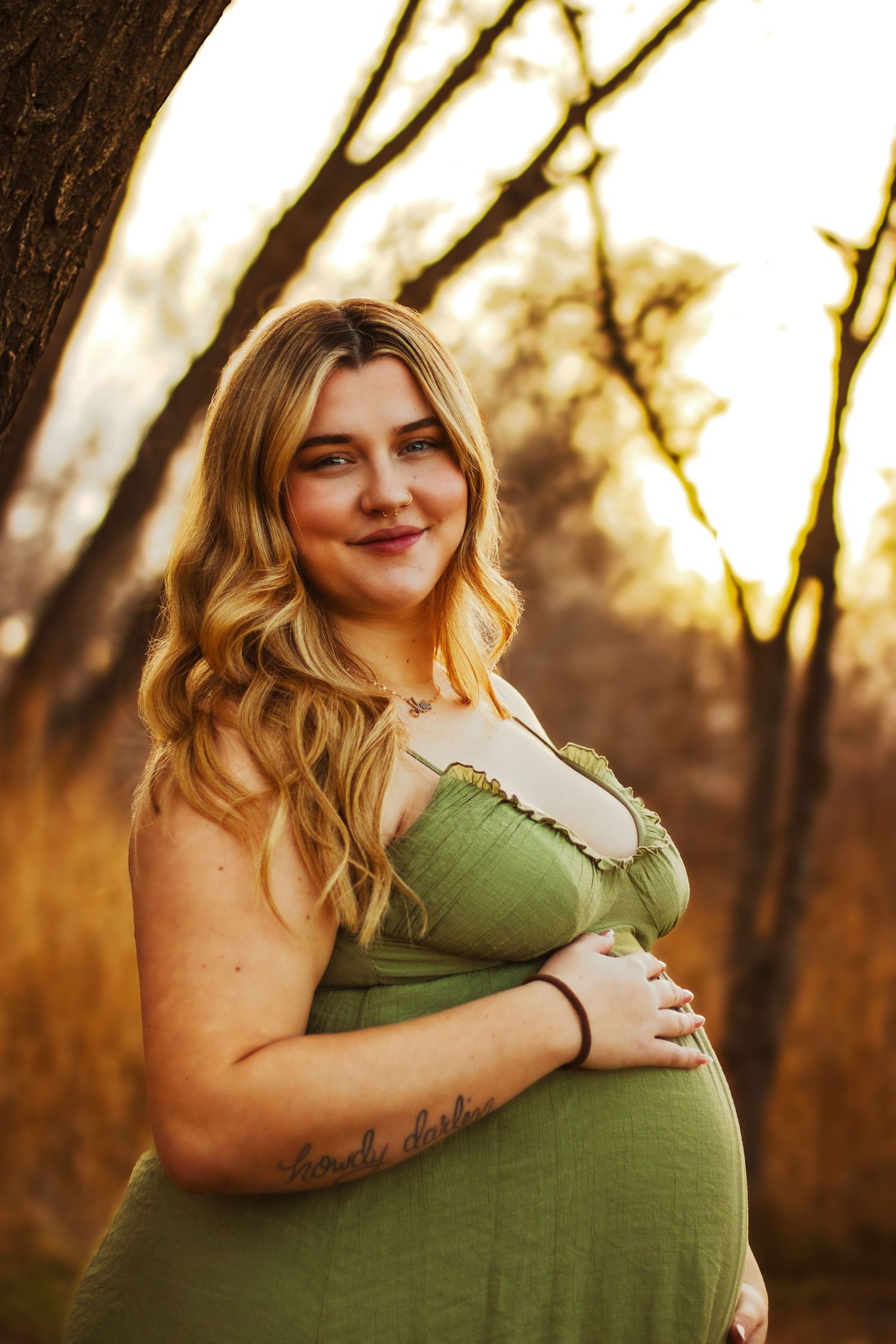 Pregnant woman in green dress holding baby bump at golden hour outdoors.