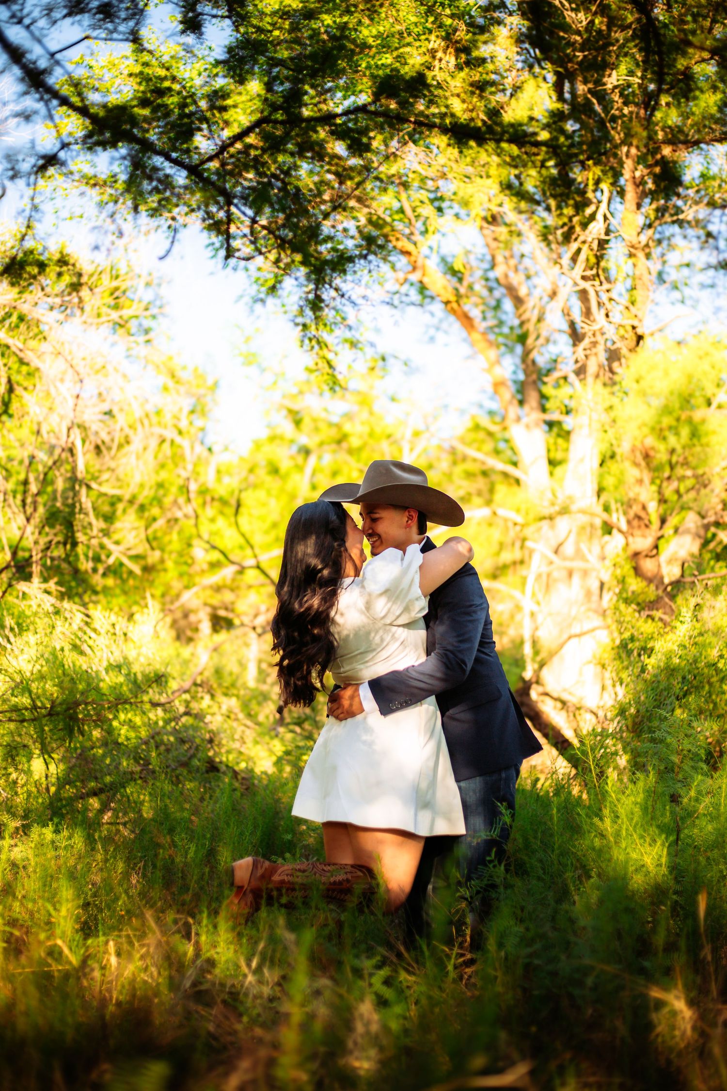 Couple kissing in a lush green forest, woman in white dress, man in navy suit and cowboy hat, golden sunlight filtering through trees.