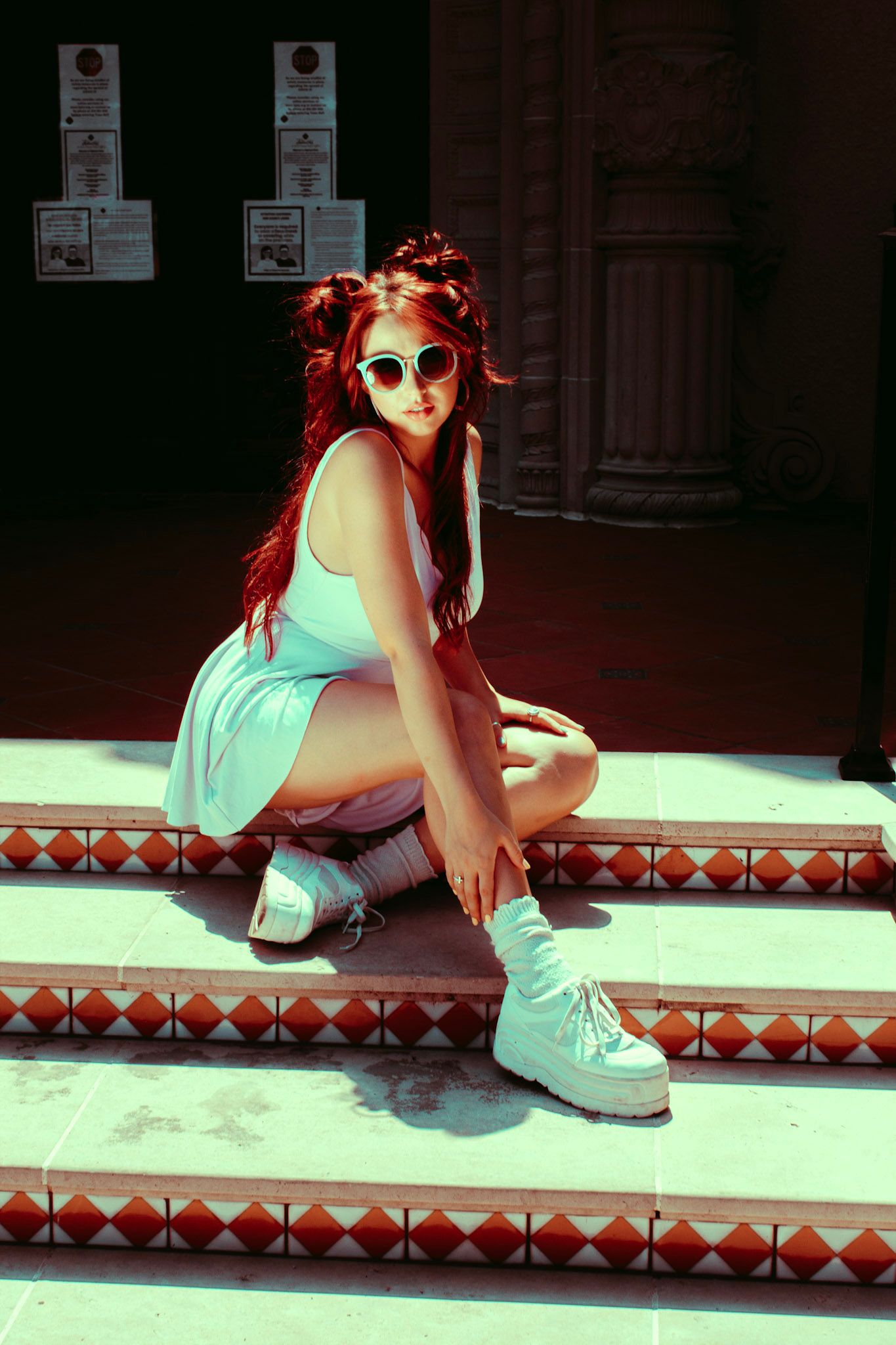 Young woman with red hair and sunglasses crouching on decorative tiled steps in sunlight.