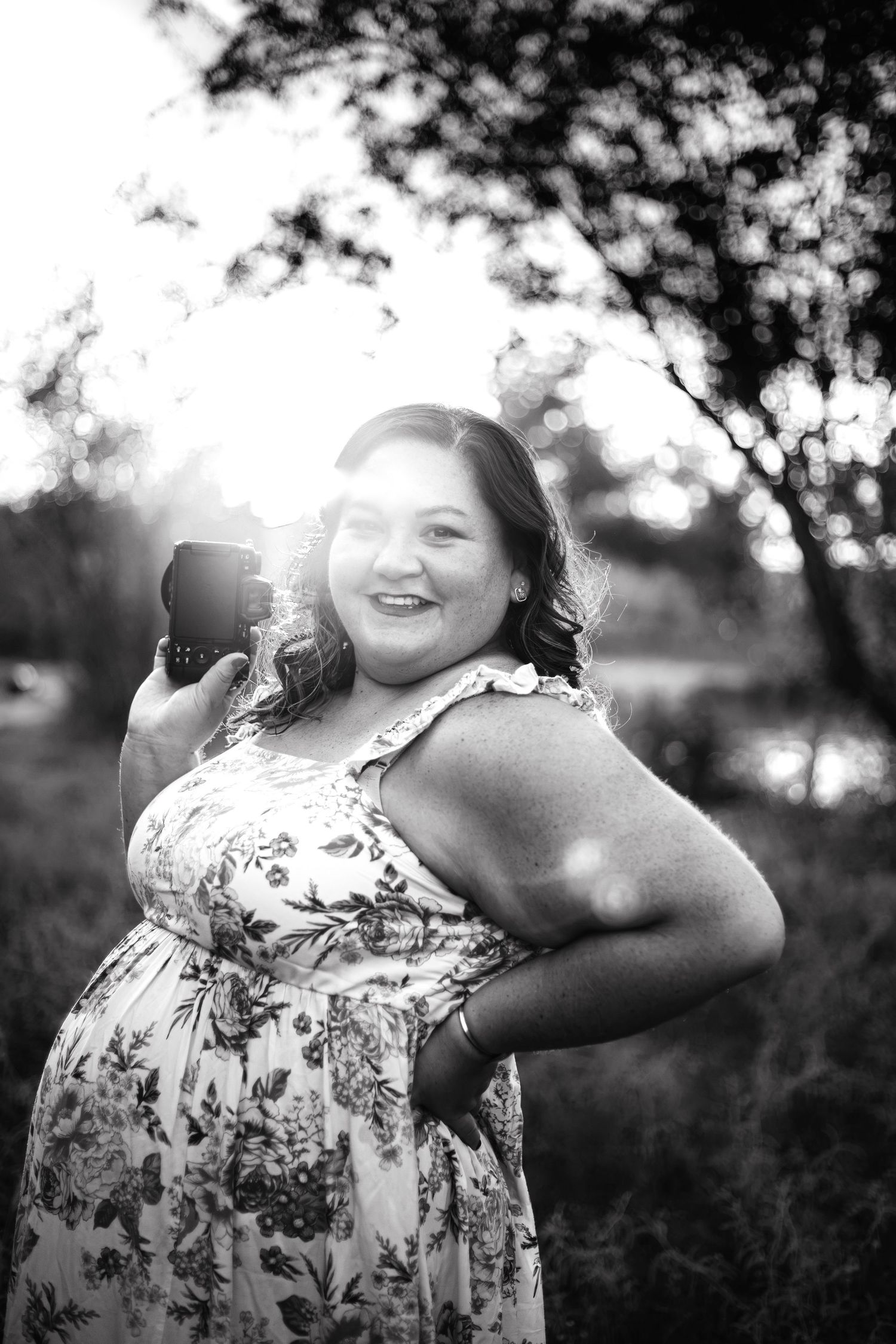 Black and white photo of a smiling woman in floral dress holding a camera outdoors with sunlight streaming through trees.