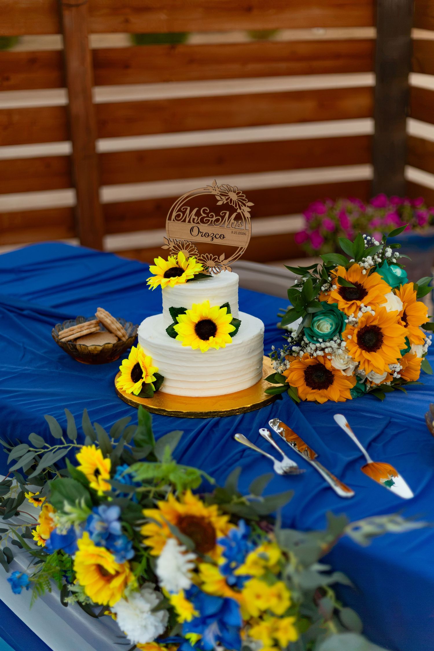 Two-tier white wedding cake with sunflowers on blue tablecloth beside sunflower bouquets outdoors.