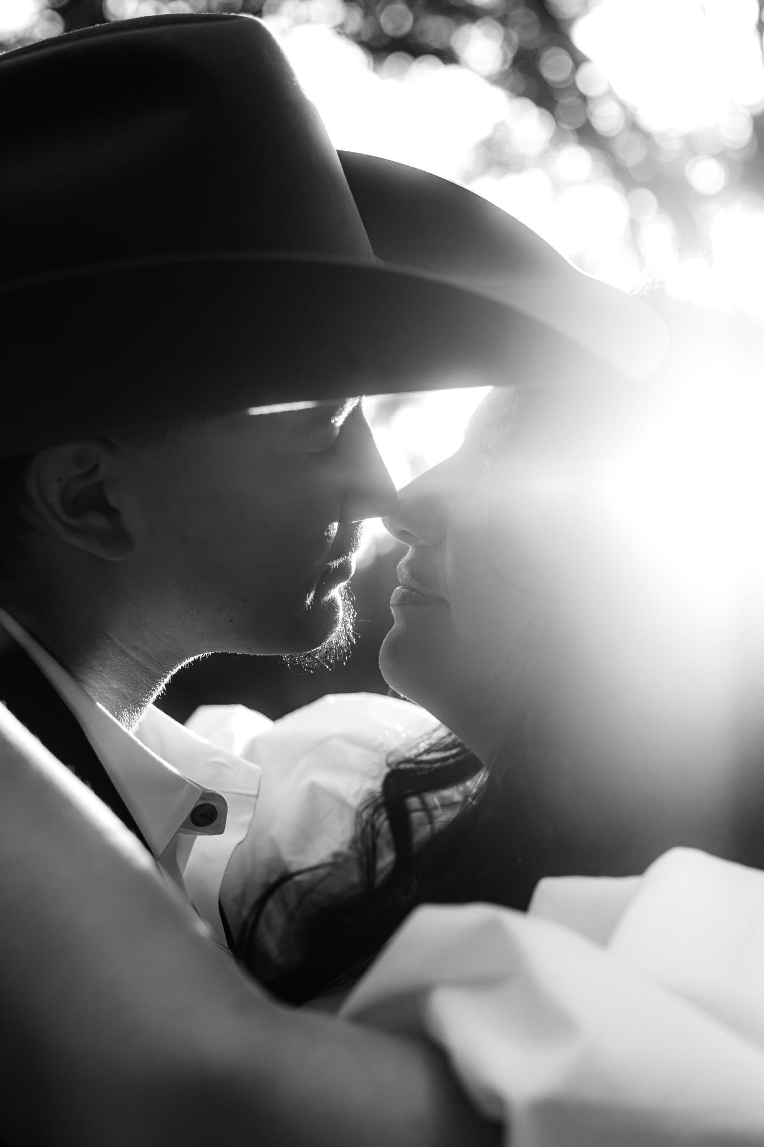 Black and white photo of a couple sharing a romantic kiss under a cowboy hat with sunlight glowing behind them.