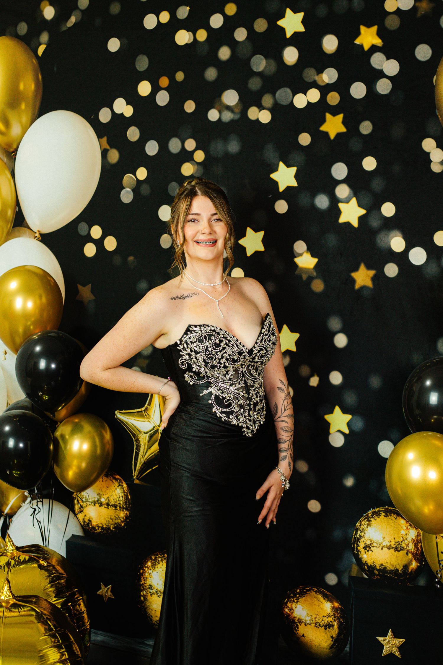 Woman in elegant black and gold gown poses with balloons and star decorations at a glamorous celebration.