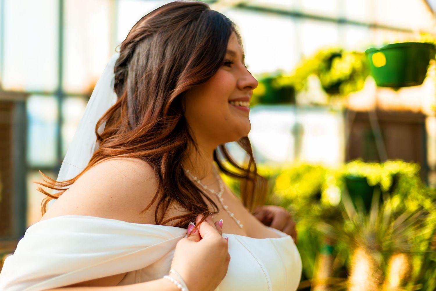 Smiling bride in white off-shoulder dress with pearl necklace, surrounded by lush green plants in warm sunlight.