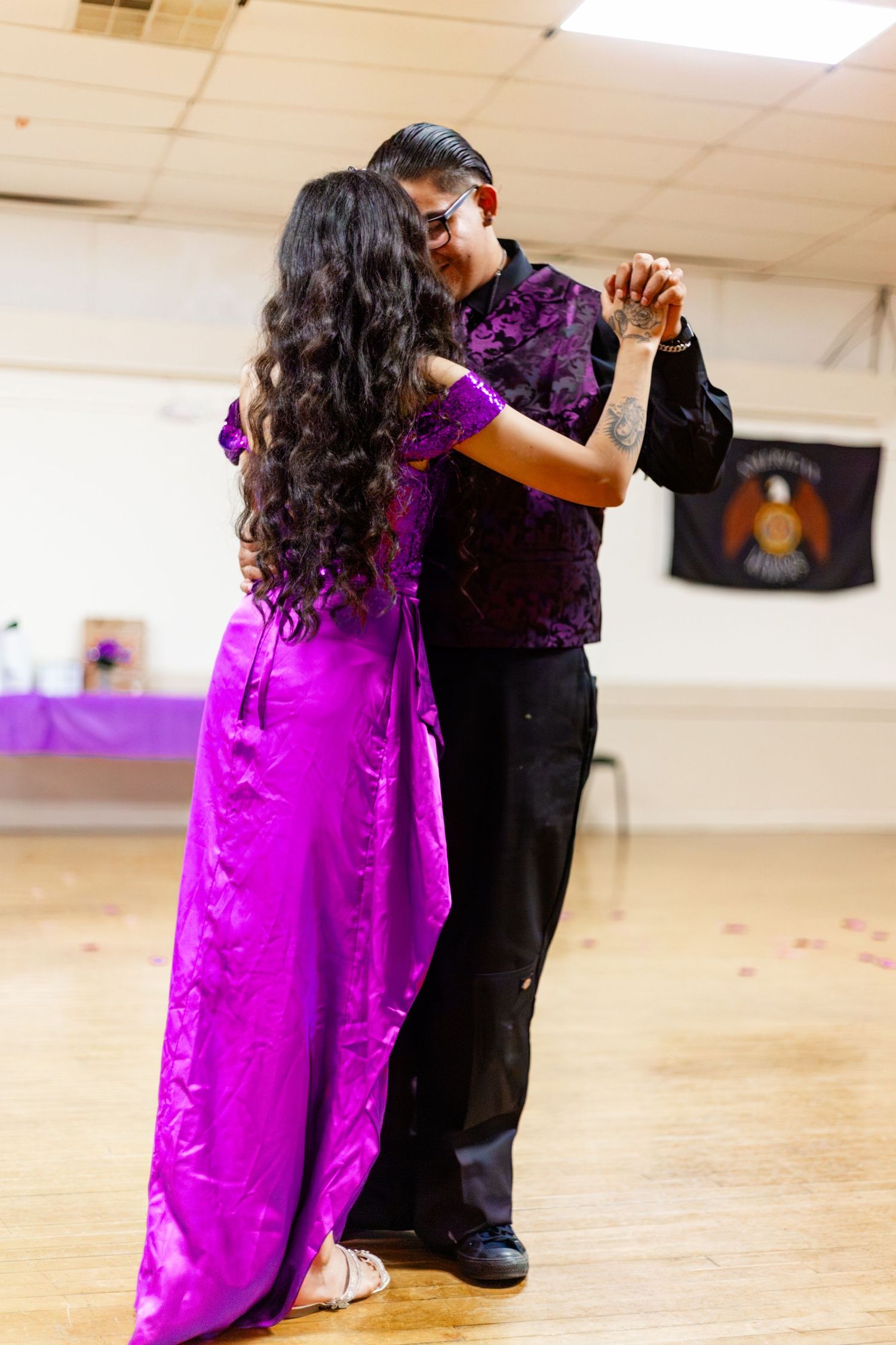 Couple dancing in formal attire, woman in purple gown, man in black shirt, on wooden dance floor.