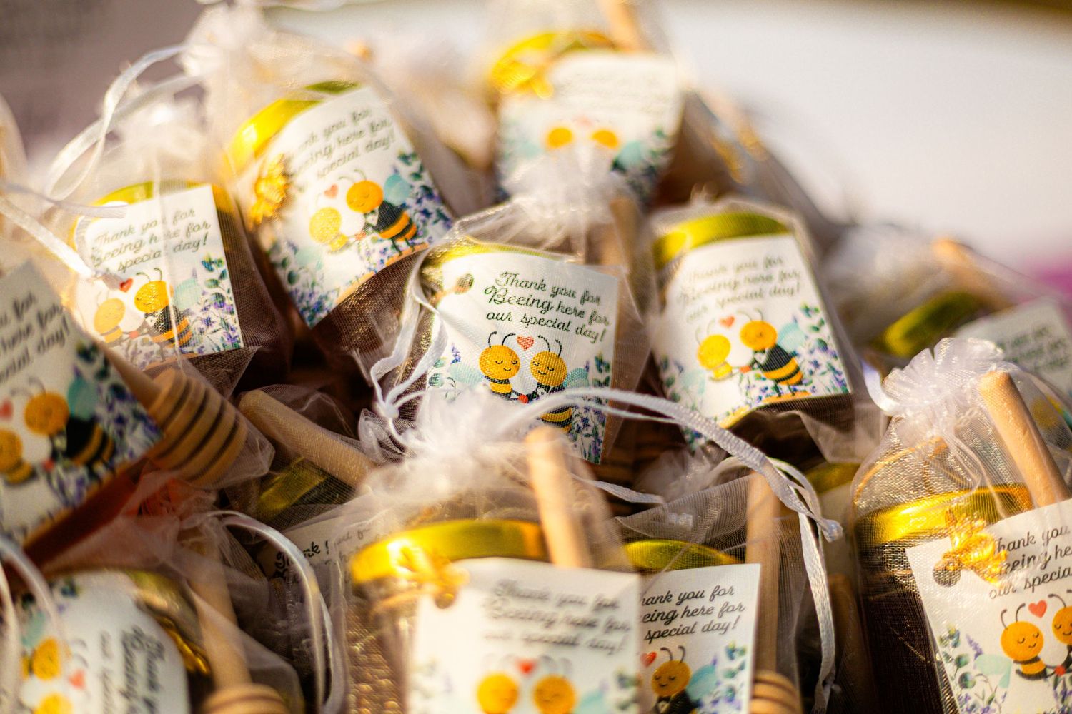 Honey jar wedding favors with bee-themed labels and wooden honey dippers wrapped in clear cellophane bags.