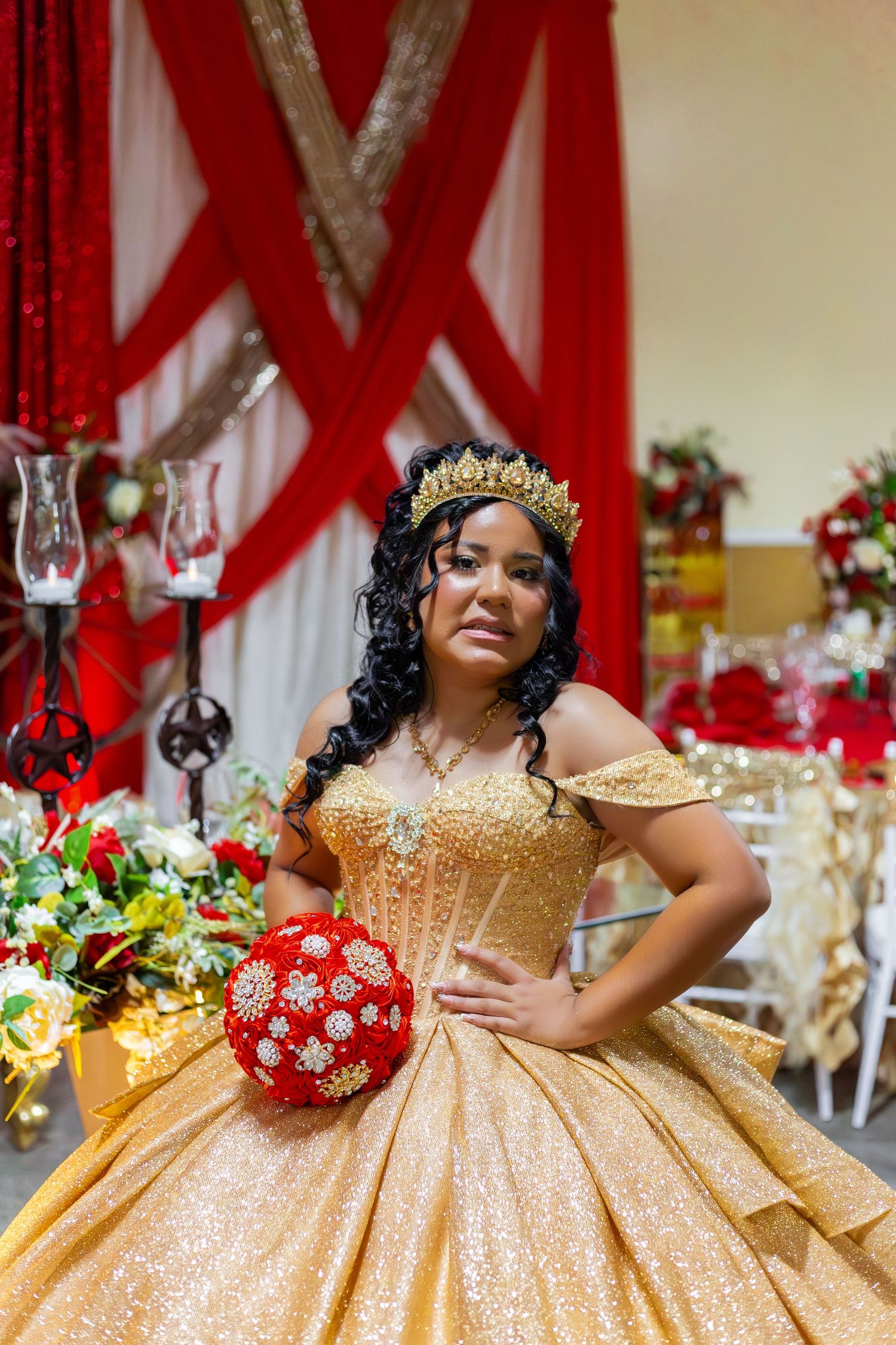 Young woman in gold quinceañera gown and crown holds red bouquet at elegant red and gold decorated venue.