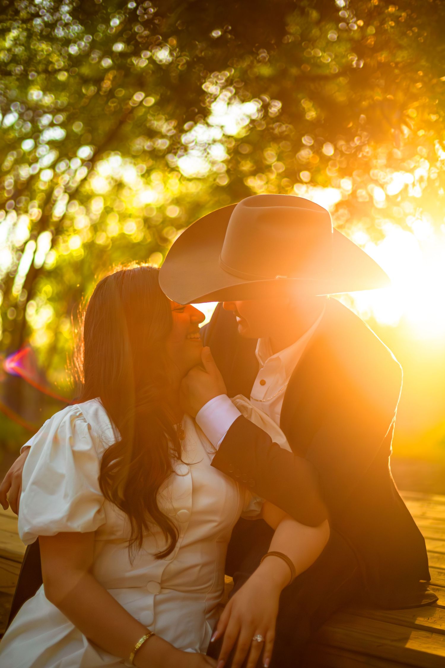 Couple sharing a romantic kiss at golden hour sunset, man wearing cowboy hat, woman in white dress, seated outdoors.