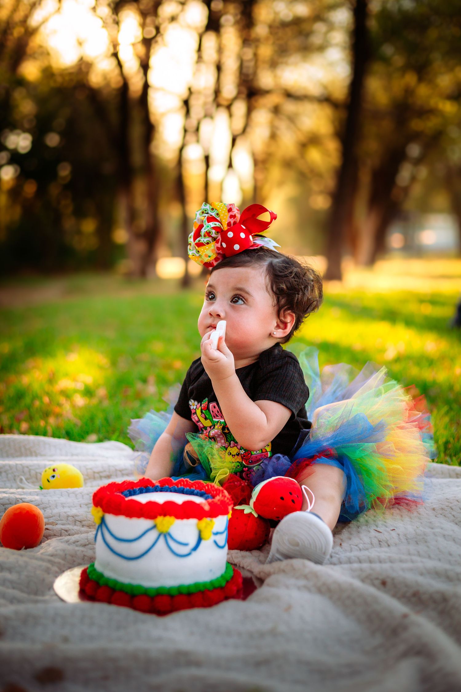 Baby girl in rainbow tutu and colorful bow celebrates first birthday with cake smash in sunny park.