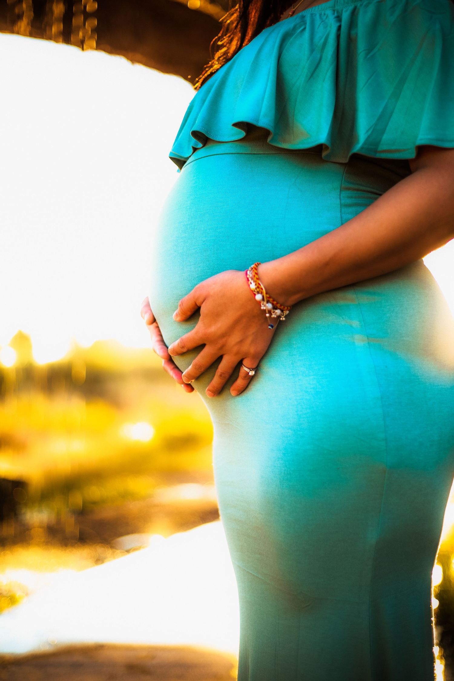Close-up of pregnant woman in teal off-shoulder dress cradling baby bump in sunlight.