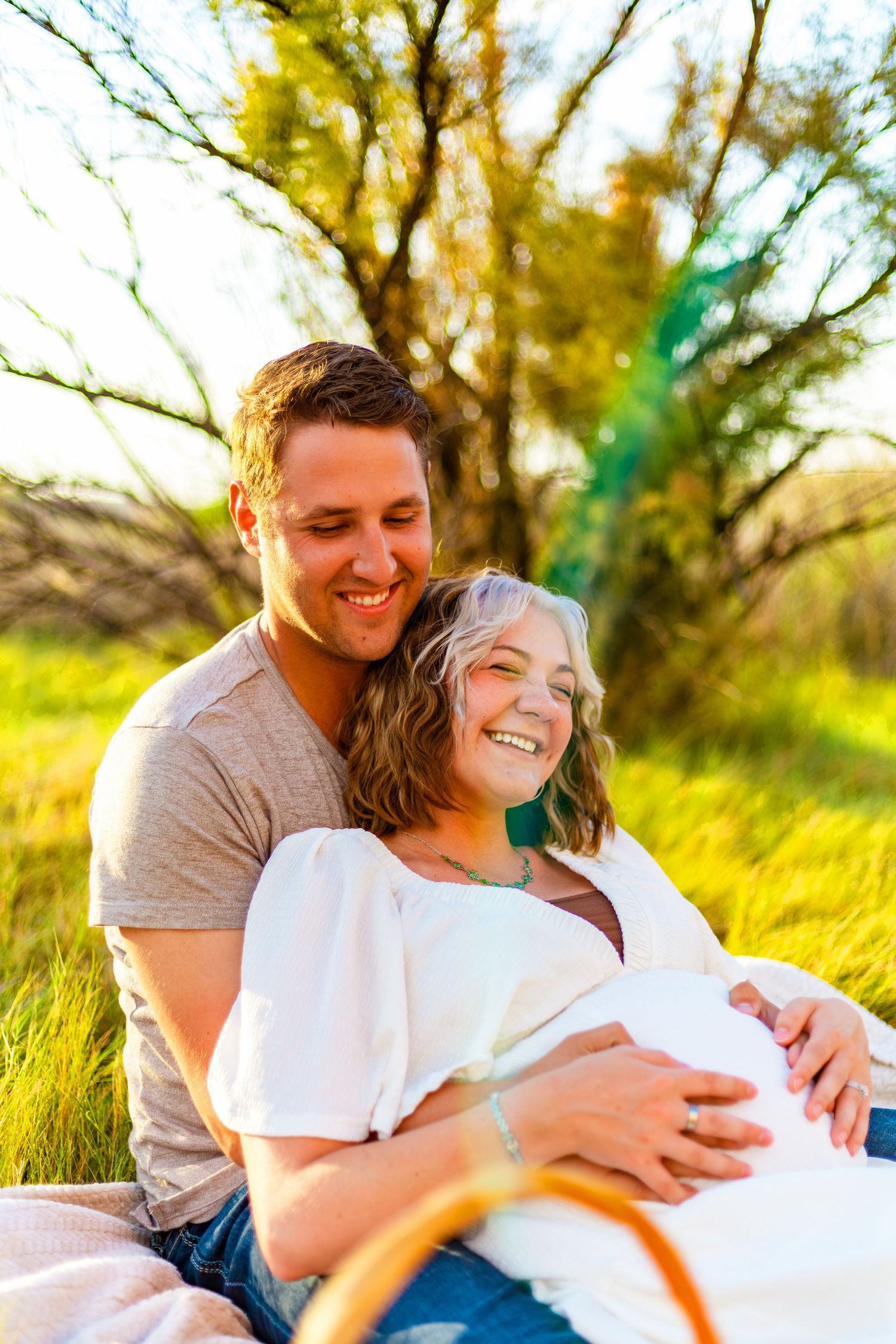 Happy couple sitting in a sunny field, pregnant woman leaning against smiling man outdoors.