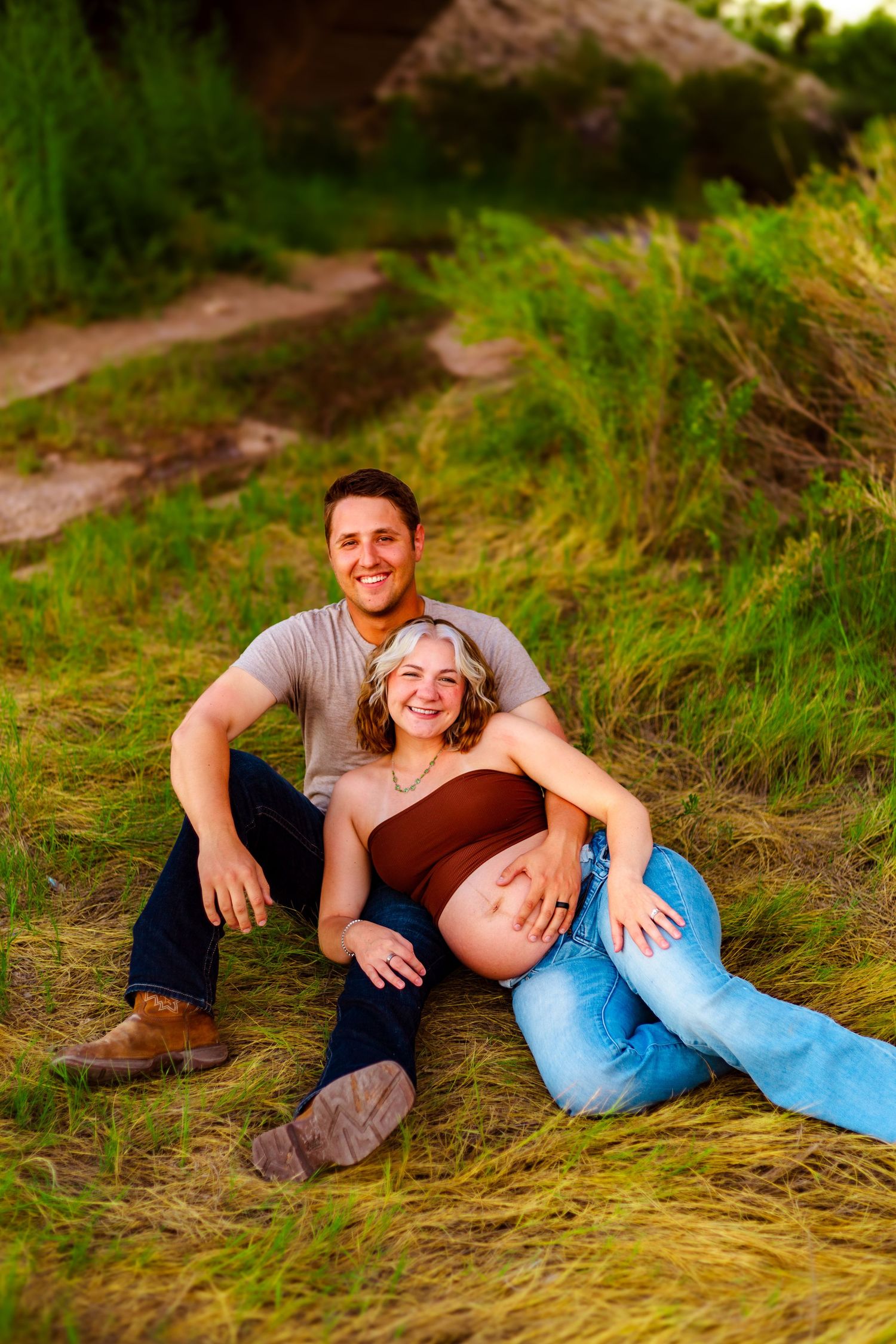 Happy couple sitting on grass, pregnant woman leaning back on man, both smiling outdoors in lush green field.