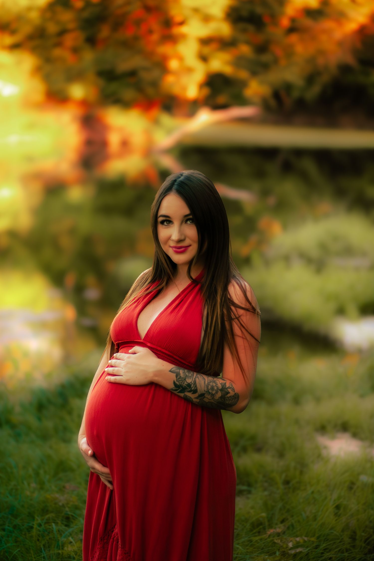 Pregnant woman in red dress holding baby bump outdoors in autumn park at golden hour.