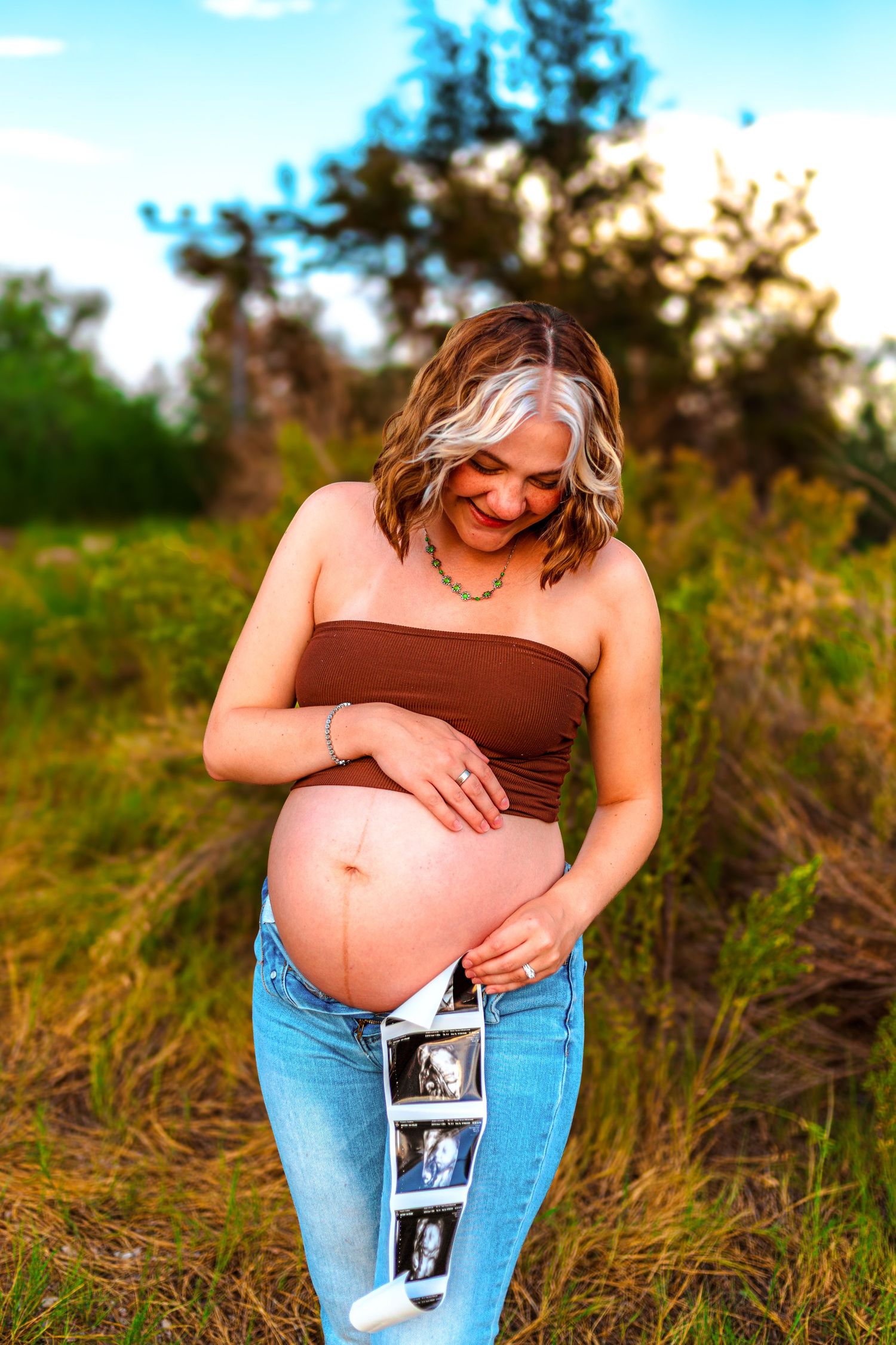 Pregnant woman in brown tube top and jeans holds ultrasound photos while cradling her baby bump outdoors at golden hour.