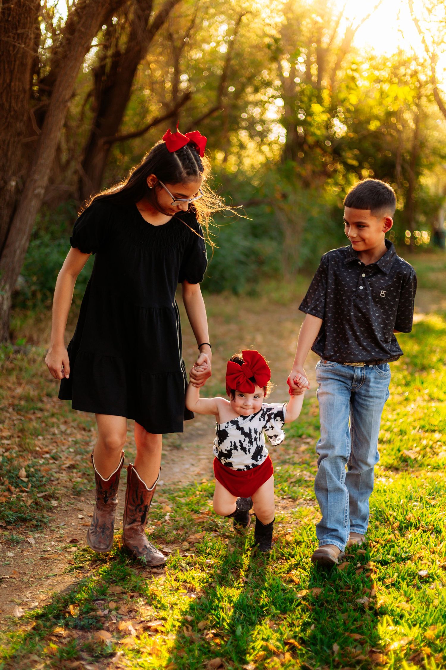 Two older siblings hold hands with a toddler in a red bow while walking outdoors in golden sunset light.