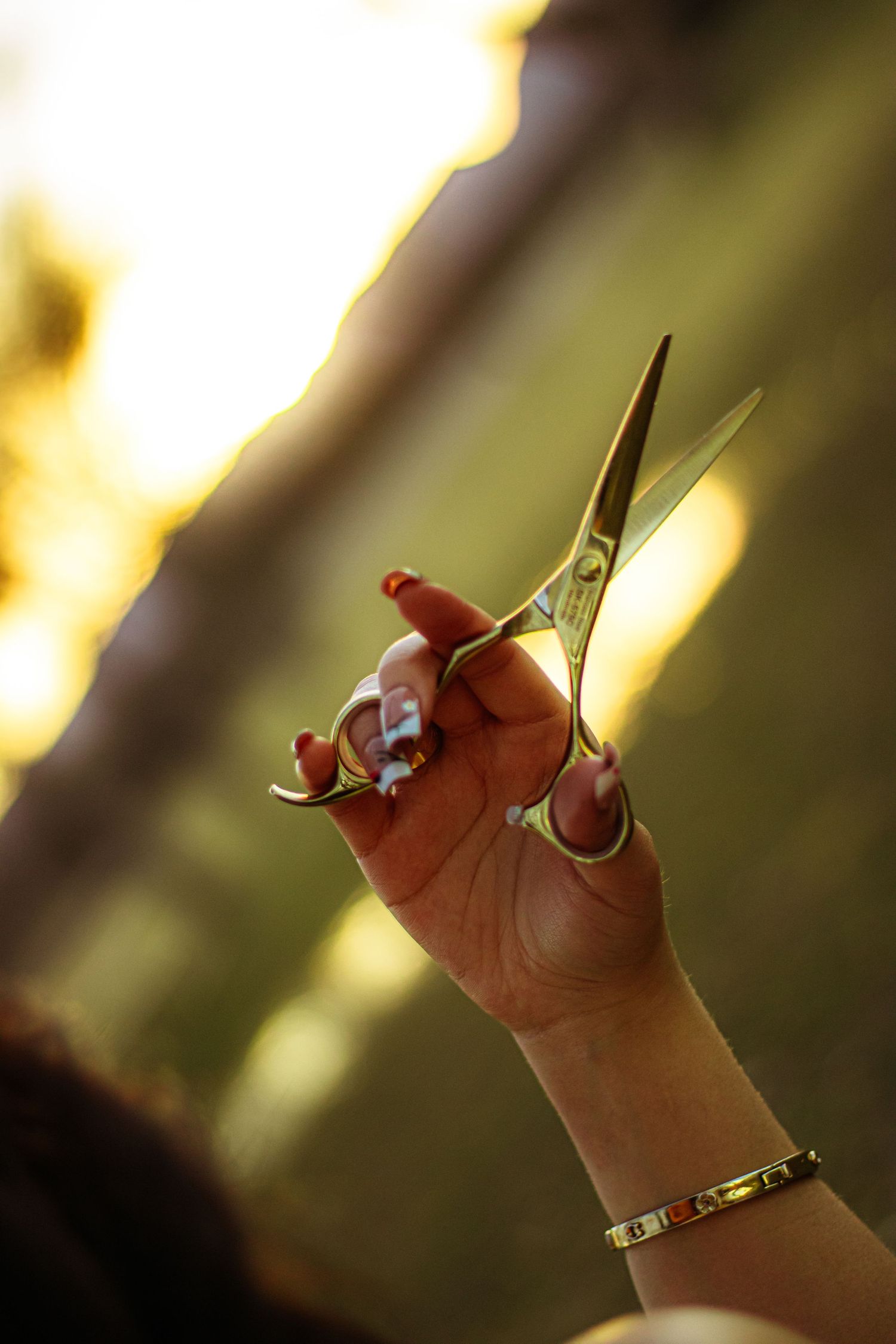 Hand holding gold hairdressing scissors outdoors in golden sunlight, wearing a bracelet with red manicured nails.