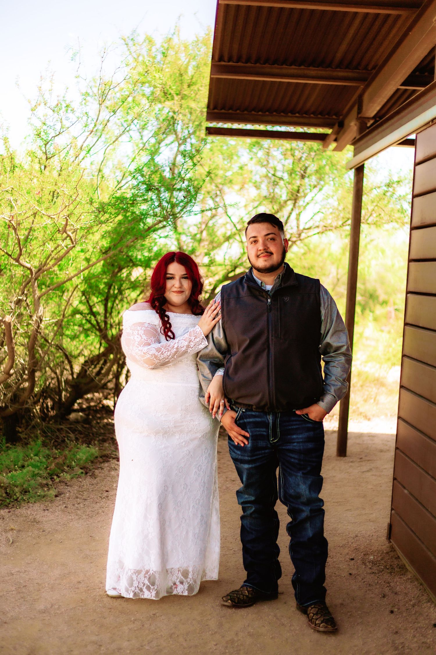 Couple posing outdoors in wedding attire, bride in white lace gown with red hair, groom in vest and jeans.