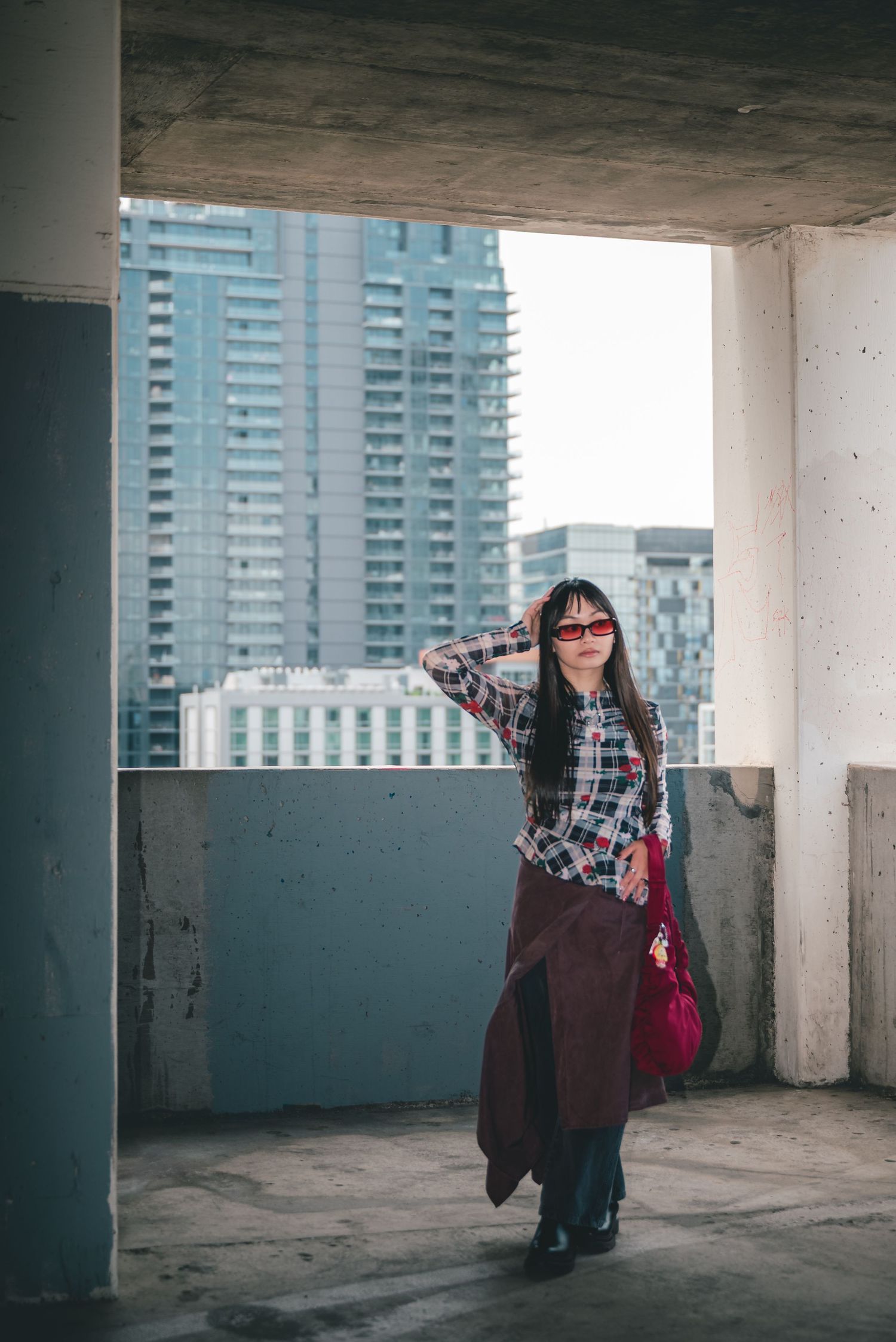 Full body portrait of a woman in business-casual fashion standing in a dirty parking garage with a view of buildings behind her.