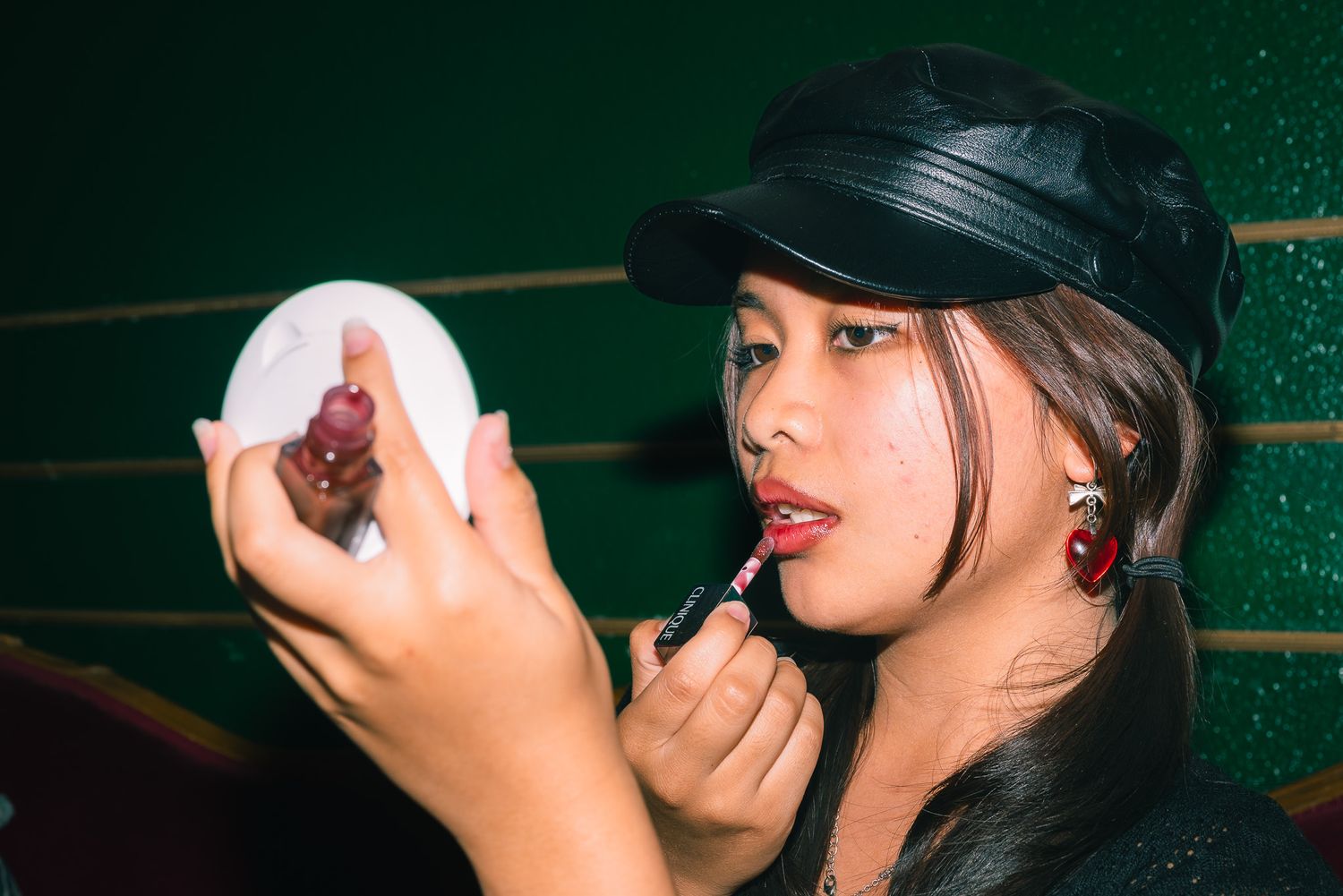 Up-close headshot of a woman in a black hat and black shirt staring into her compact mirror as she applies her lipstick.