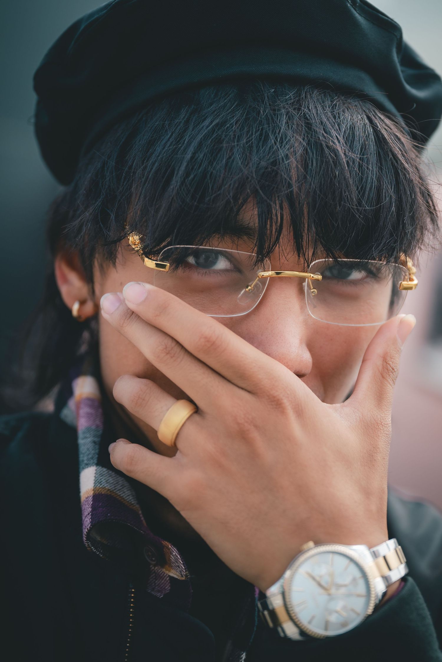 Headshot of a fashionable man in gold glasses obscuring his face with his left hand, which is adorned with a watch and engagement ring.