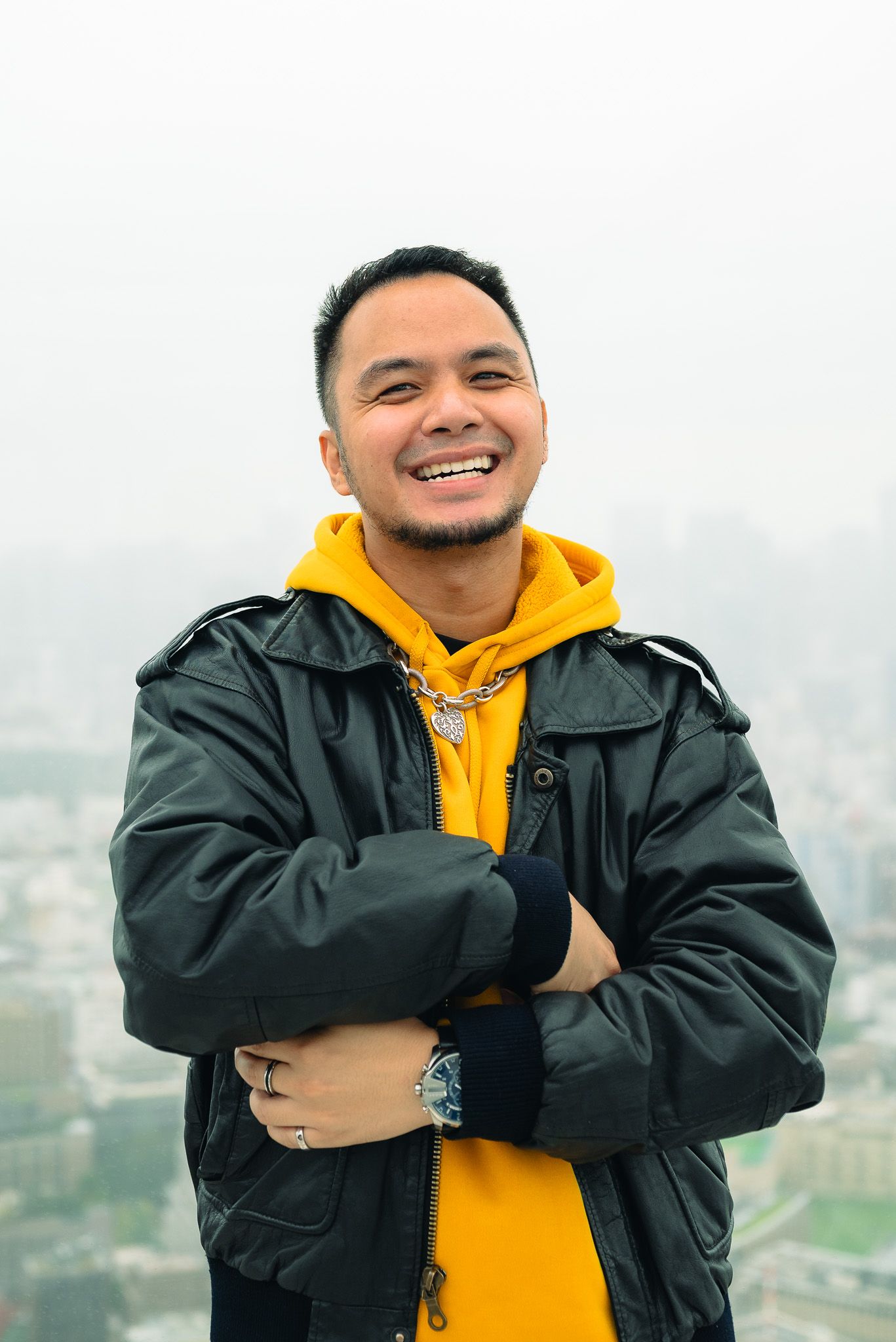 Portrait of a man in a yellow hoodie and black leather jacket crossing his arms and smiling with a cloudy view of Shibuya behind him.
