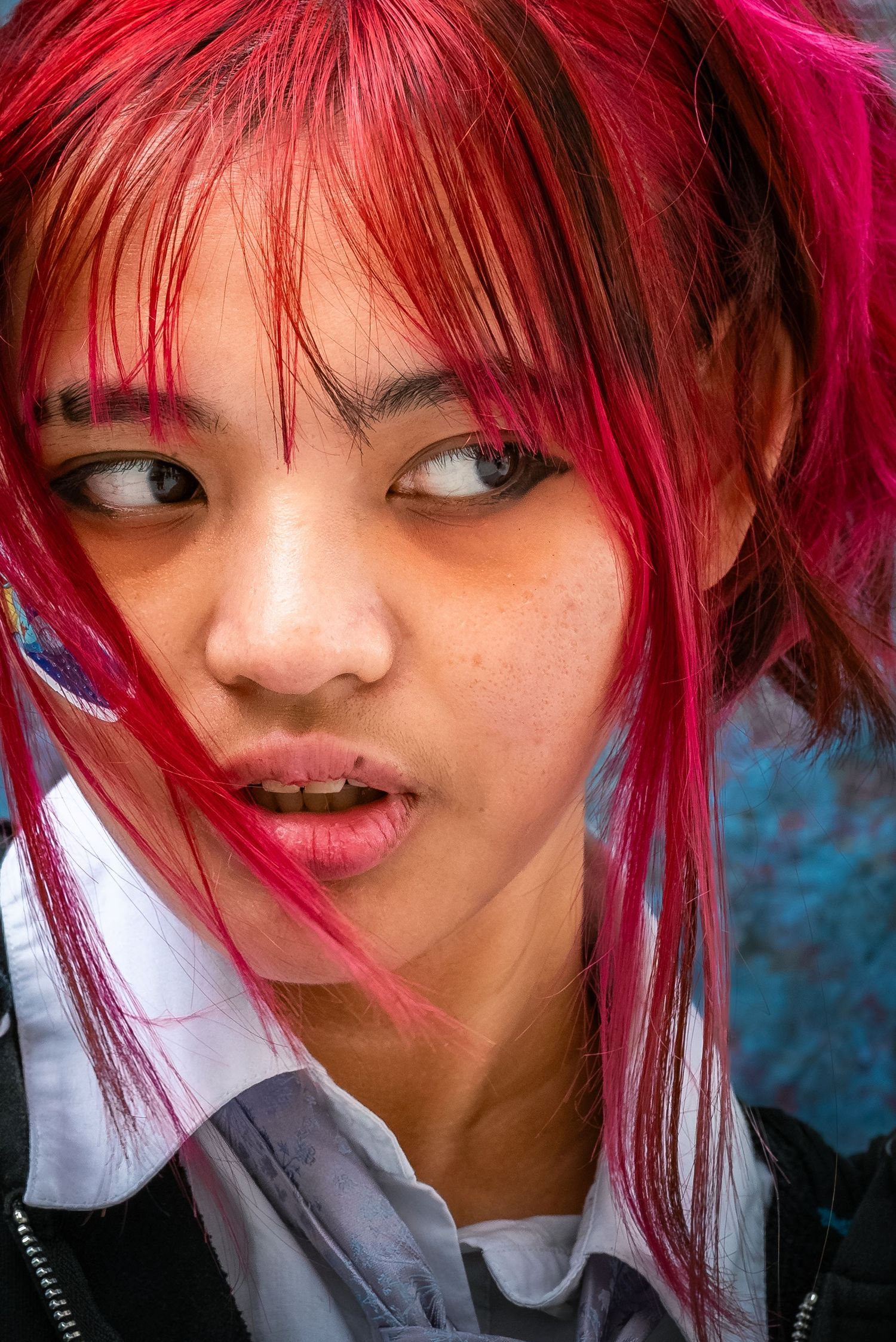 Up-close headshot of a girl with bright red hair in a school uniform posed in front of a blue yearbook backdrop, glaring to her left.