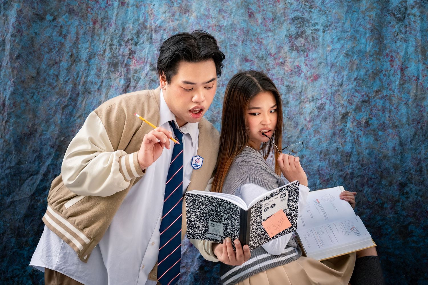 Portrait of a man and woman in school uniforms in front of a blue yearbook backdrop, both looking down at a notebook in confusion