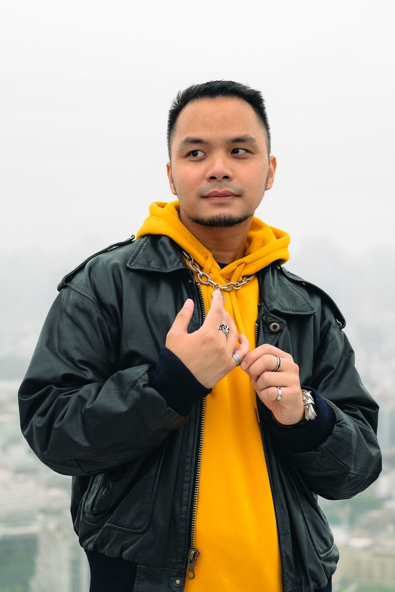 Portrait of a man in a yellow hoodie and black leather jacket fiddling with his rings with a cloudy view of Shibuya behind him.