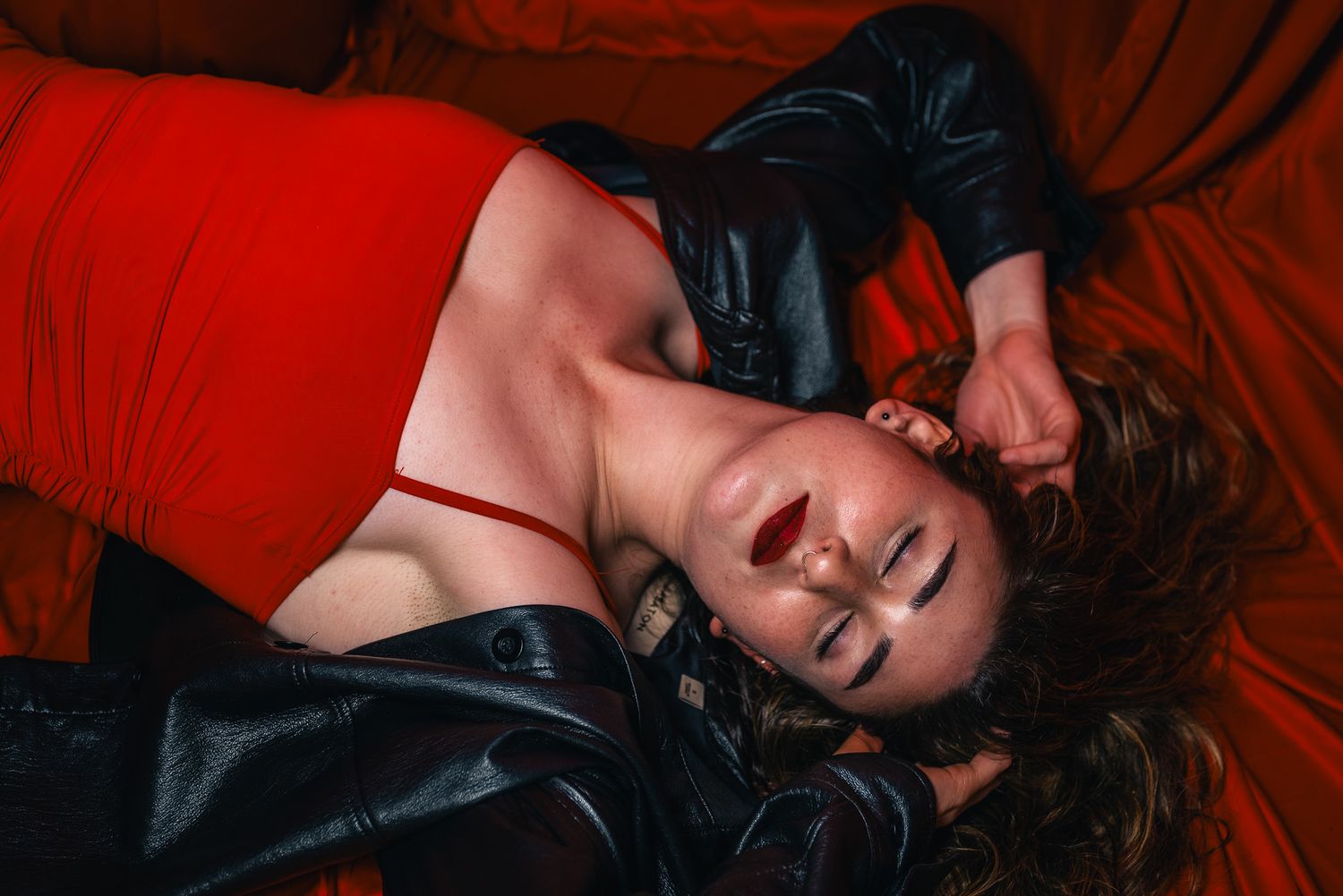 Portrait of a woman in a red dress and black leather jacket laying down with her arms behind her head, as if dreaming.