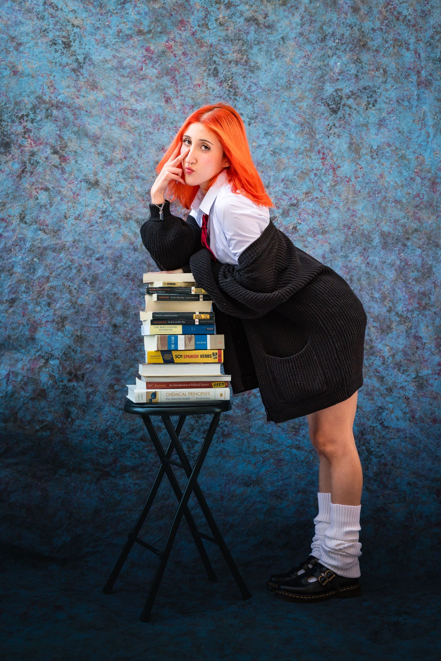 Full-body portrait of a girl with orange hair in a school uniform leaning on a stack of books in front of a blue yearbook backdrop.