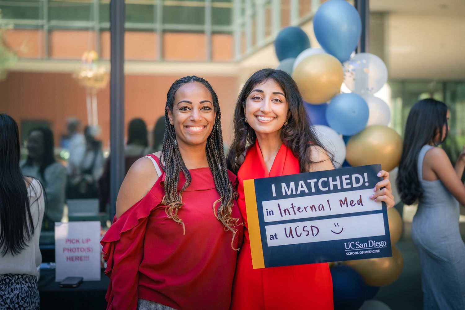 Portrait of a woman in a red dress stands proudly by her advisor, holding a blue sign celebrating her match for residency.