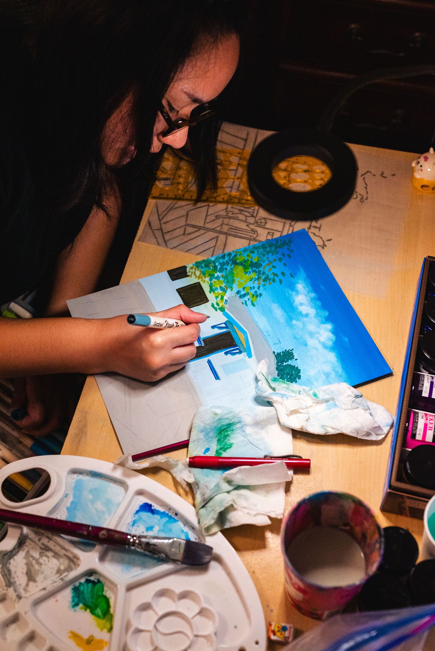 Behind the scenes shot of a woman adding detail work to her painting, her desk littered with art supplies.