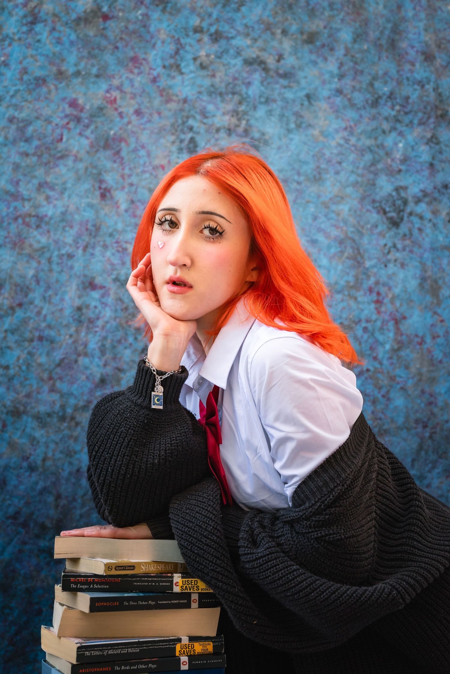 Portrait of a girl with bright orange in a school uniform in front of a blue yearbook backdrop, leaning on a stack of books.