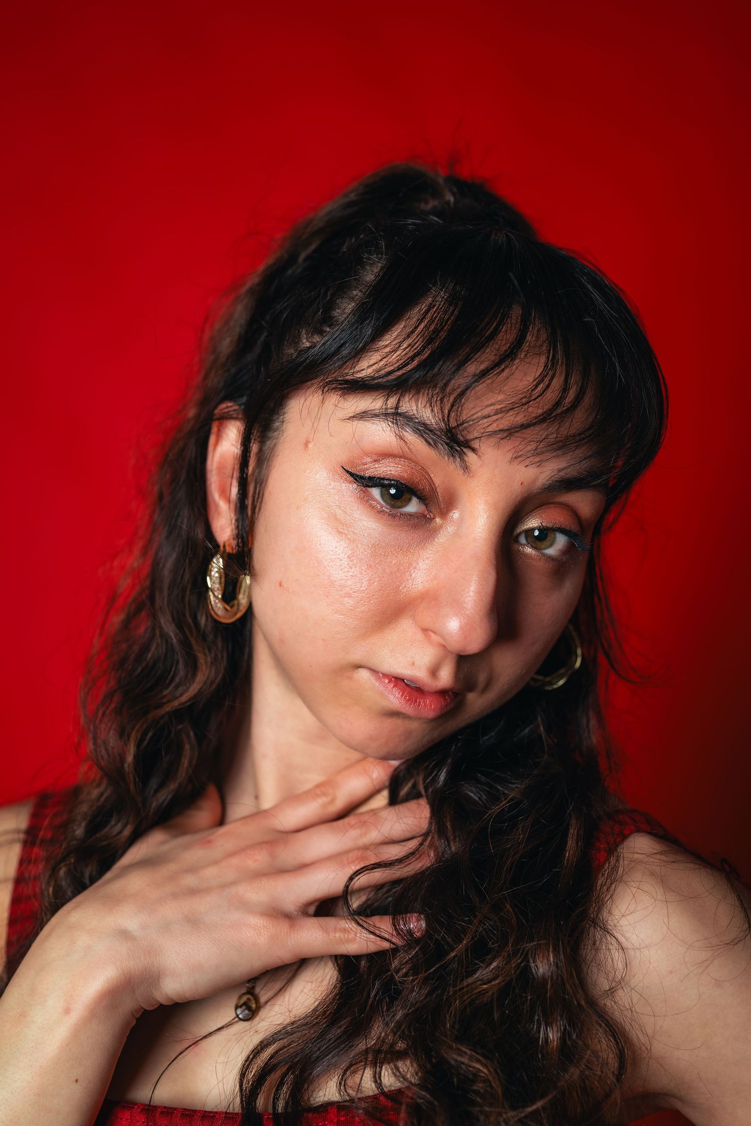 Up-close headshot of a woman in red in front of a red studio backdrop, tilting her head to the left with her right hand on her chest.
