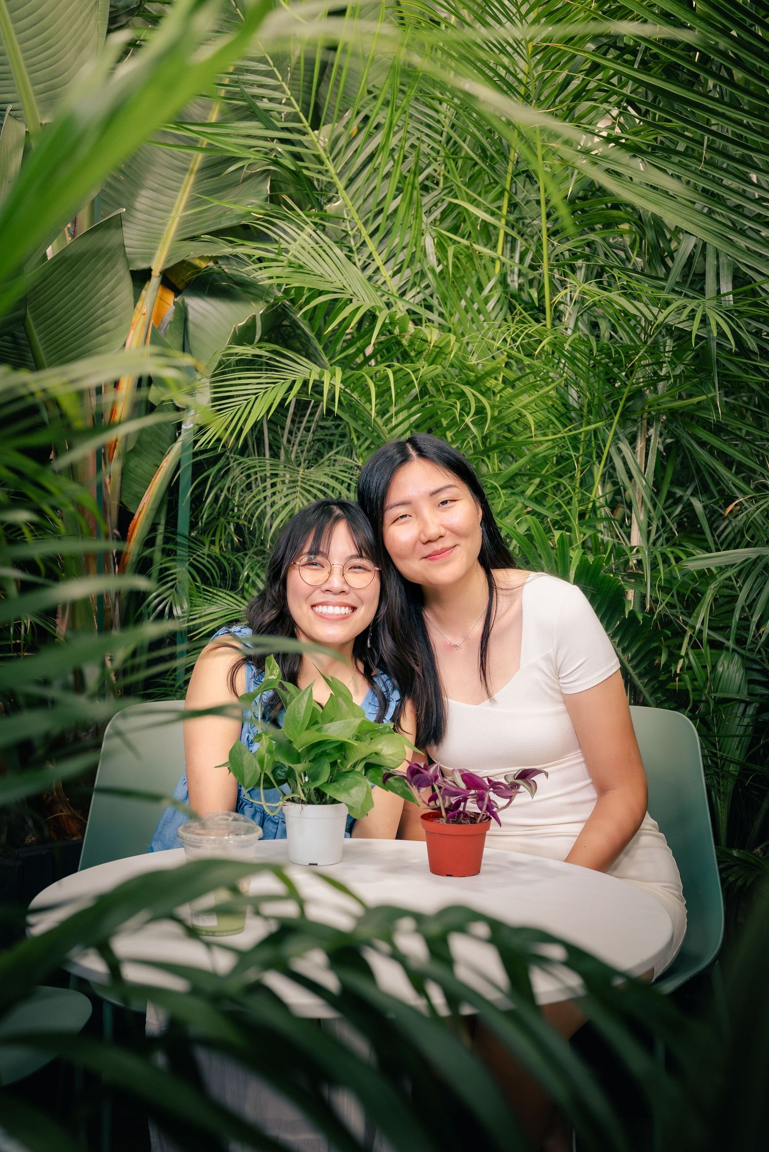 Portrait of two close friends sitting down at a white table surrounded by ample foliage, their heads leaning on each other's.