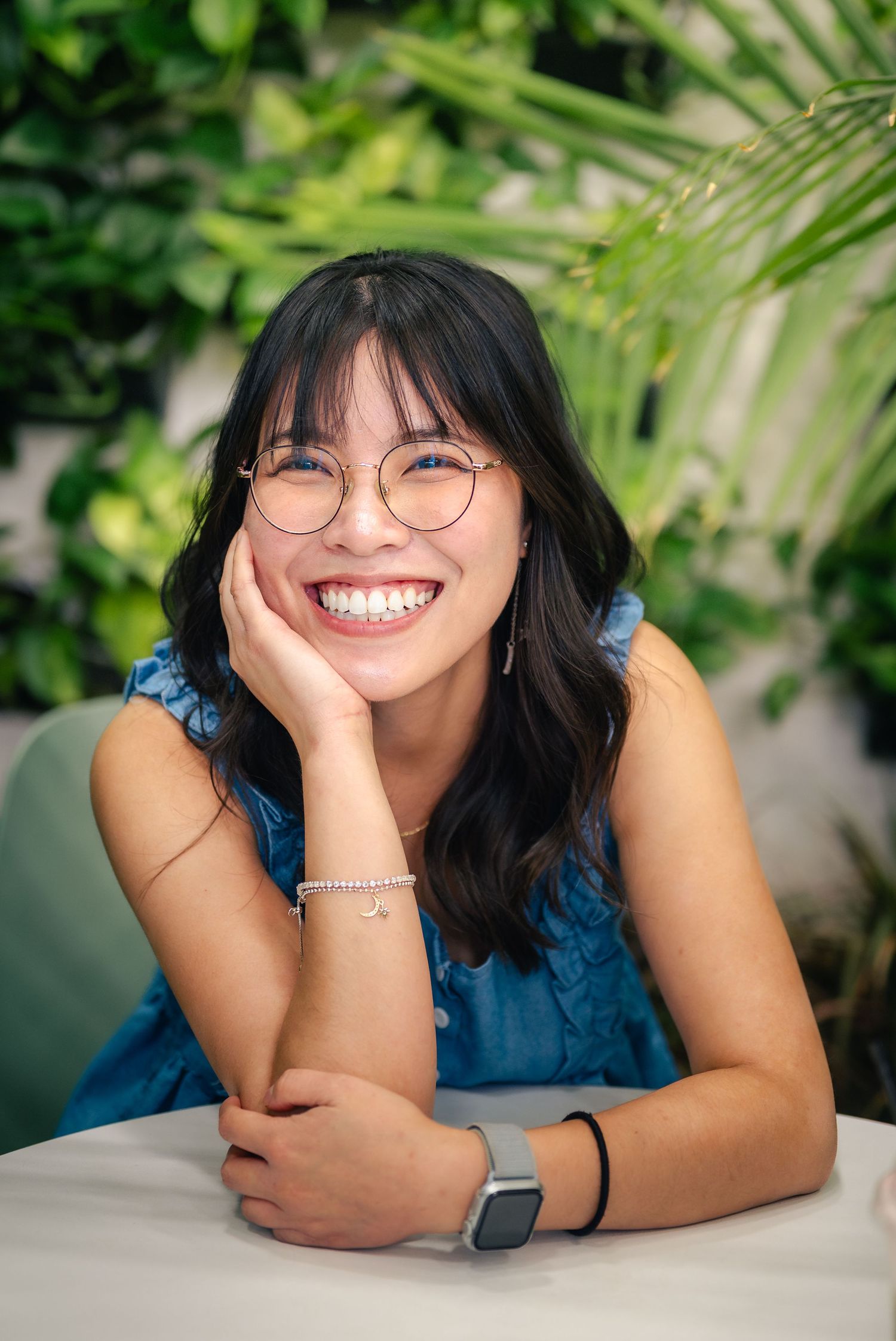 Portrait of a woman leaning sitting on a white table surrounded by foliage, smiling as she leans her cheek into her right hand.
