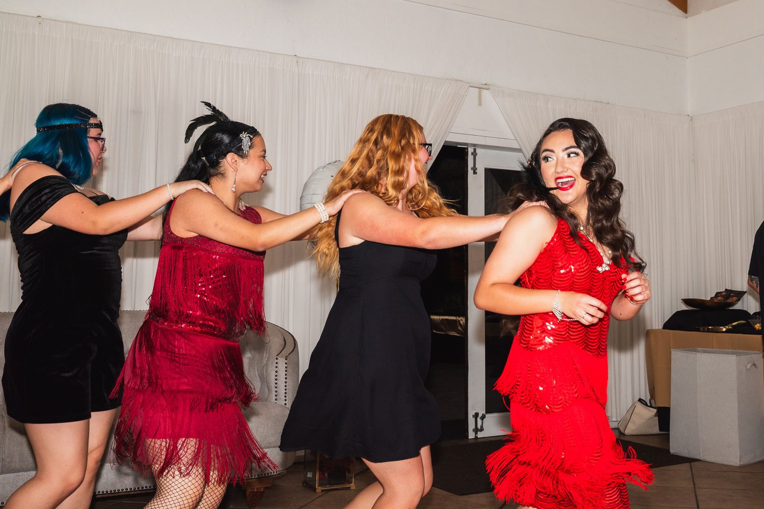 Portrait of a woman in a red dress looking back at her friends as they dance and follow her in a conga line at a 1920s-themed party.