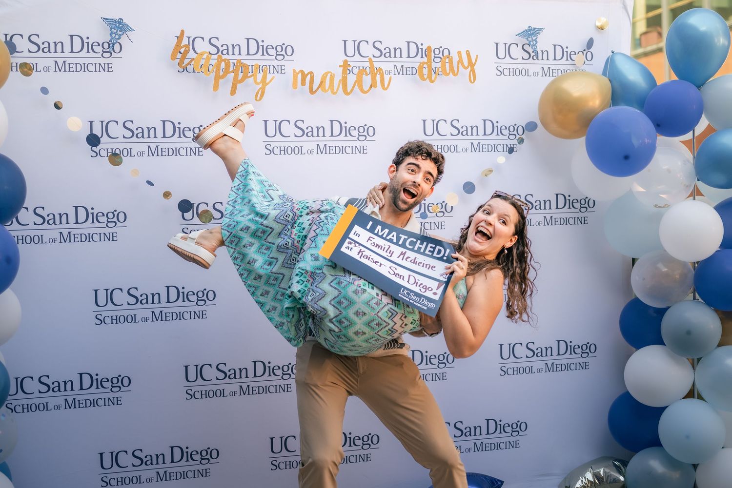 Portrait of a woman being held by her partner as she display a sign in celebration of her matching into residency.