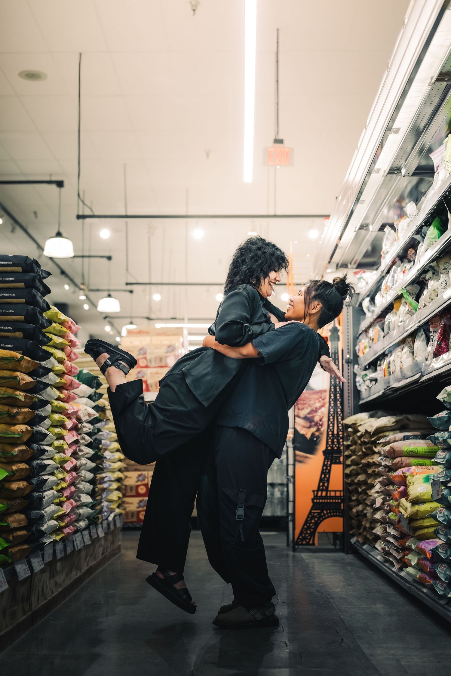 Portrait of two women in the rice aisle of HMART, one of which holds up the other by the waist as they look lovingly into each others' eyes.