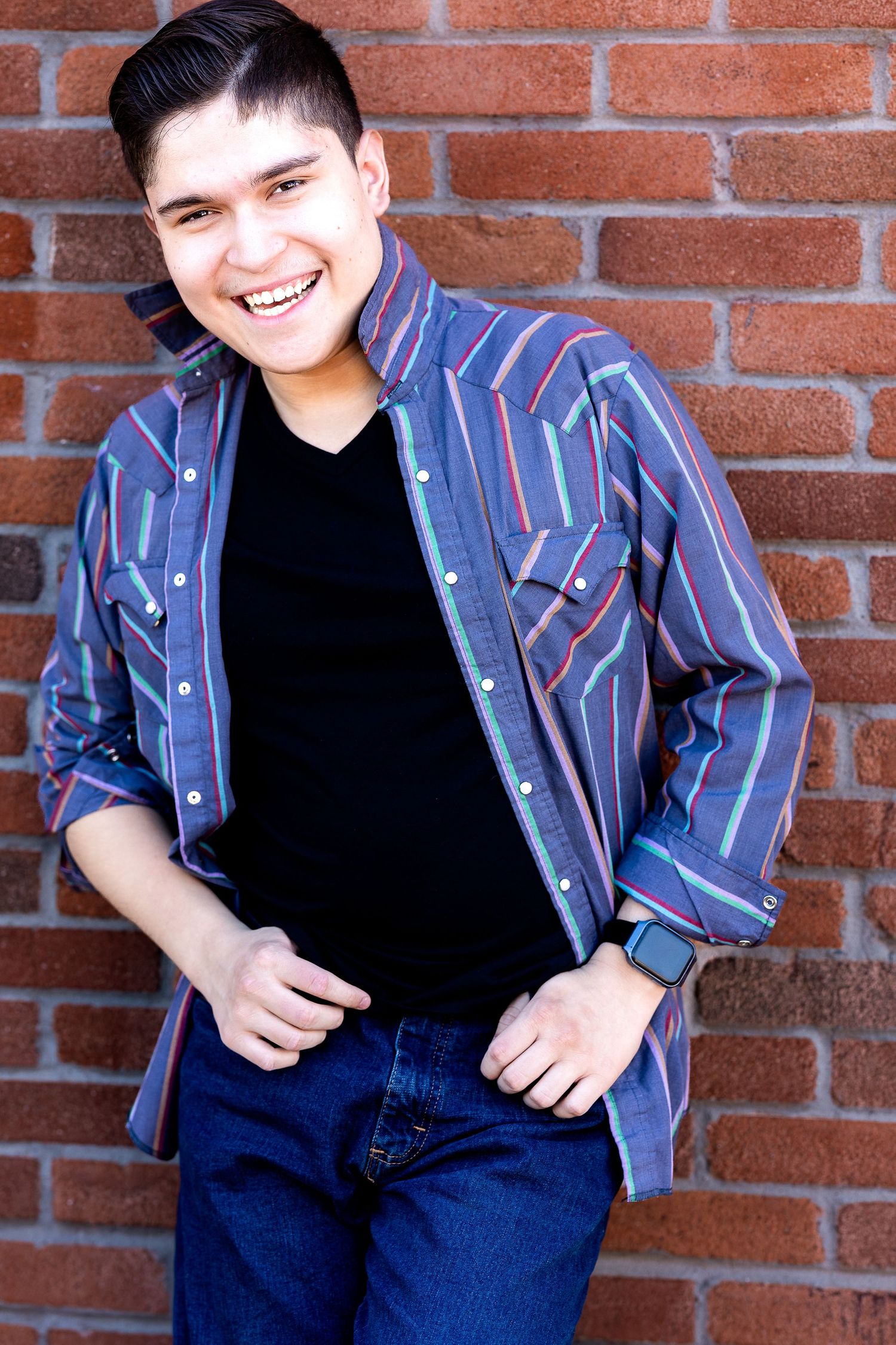 Hispanic male during his senior pictures leaning against a red brick wall in a James Dean pose.
