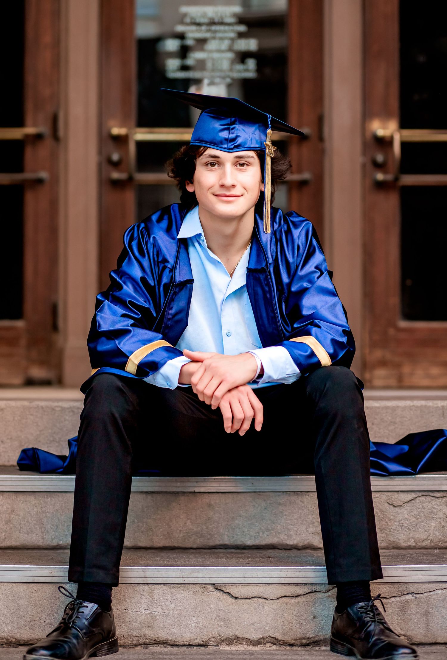 Male Senior during his Senior portrait session wearing his cap and gown, sitting on steps to a building in a downtown environment.