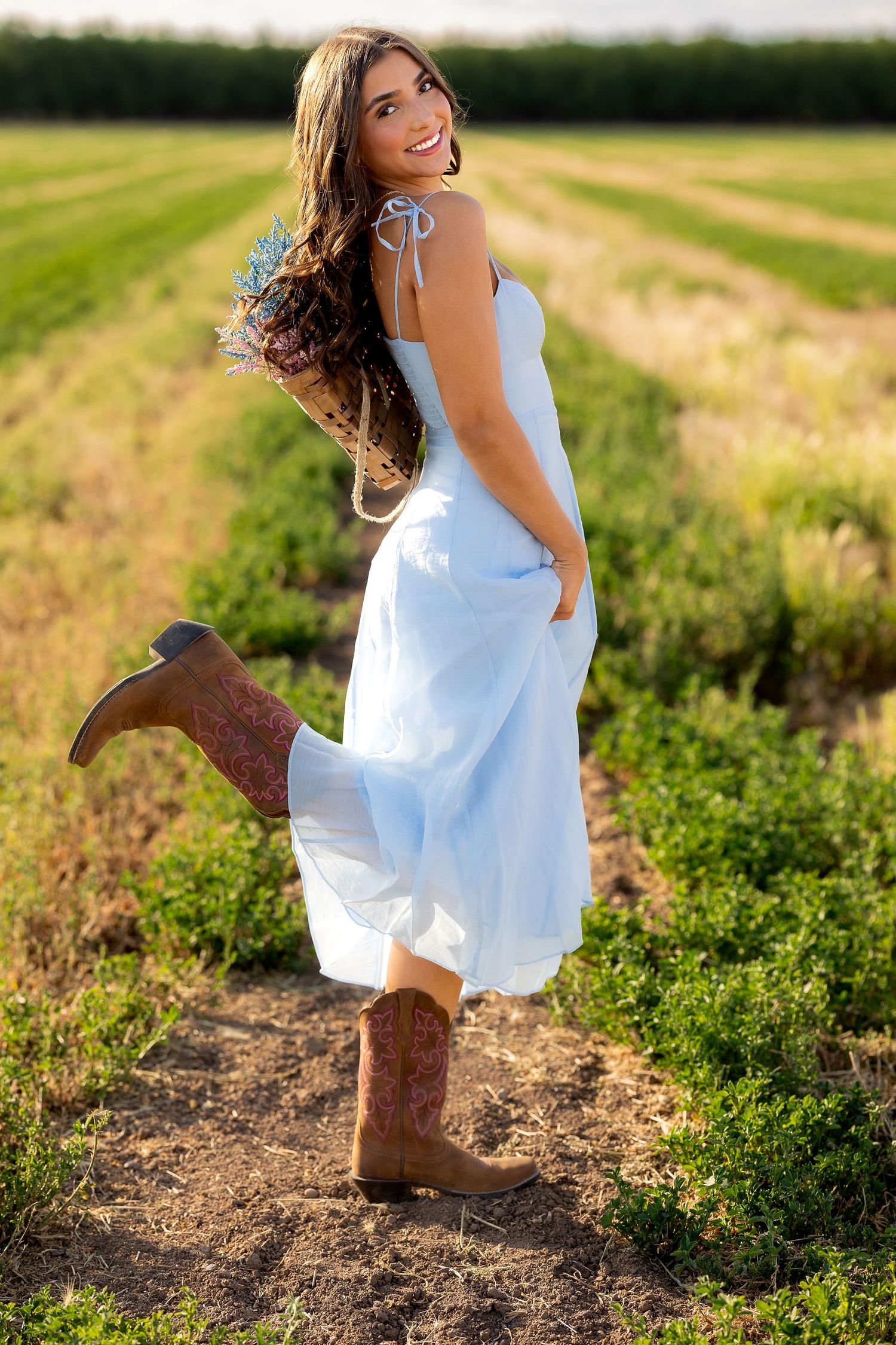 Female high School senior in soft blue dress and cowboy boots in Missouri field with floral bouquet backpack.