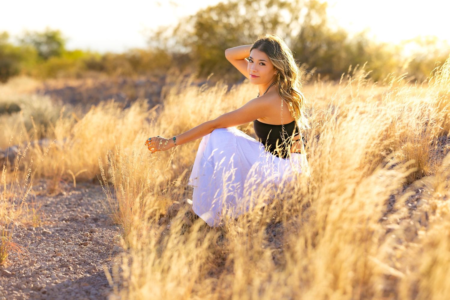 High school senior female during Senior Portraits sitting amongst tall golden grass with sun shining in her hair.