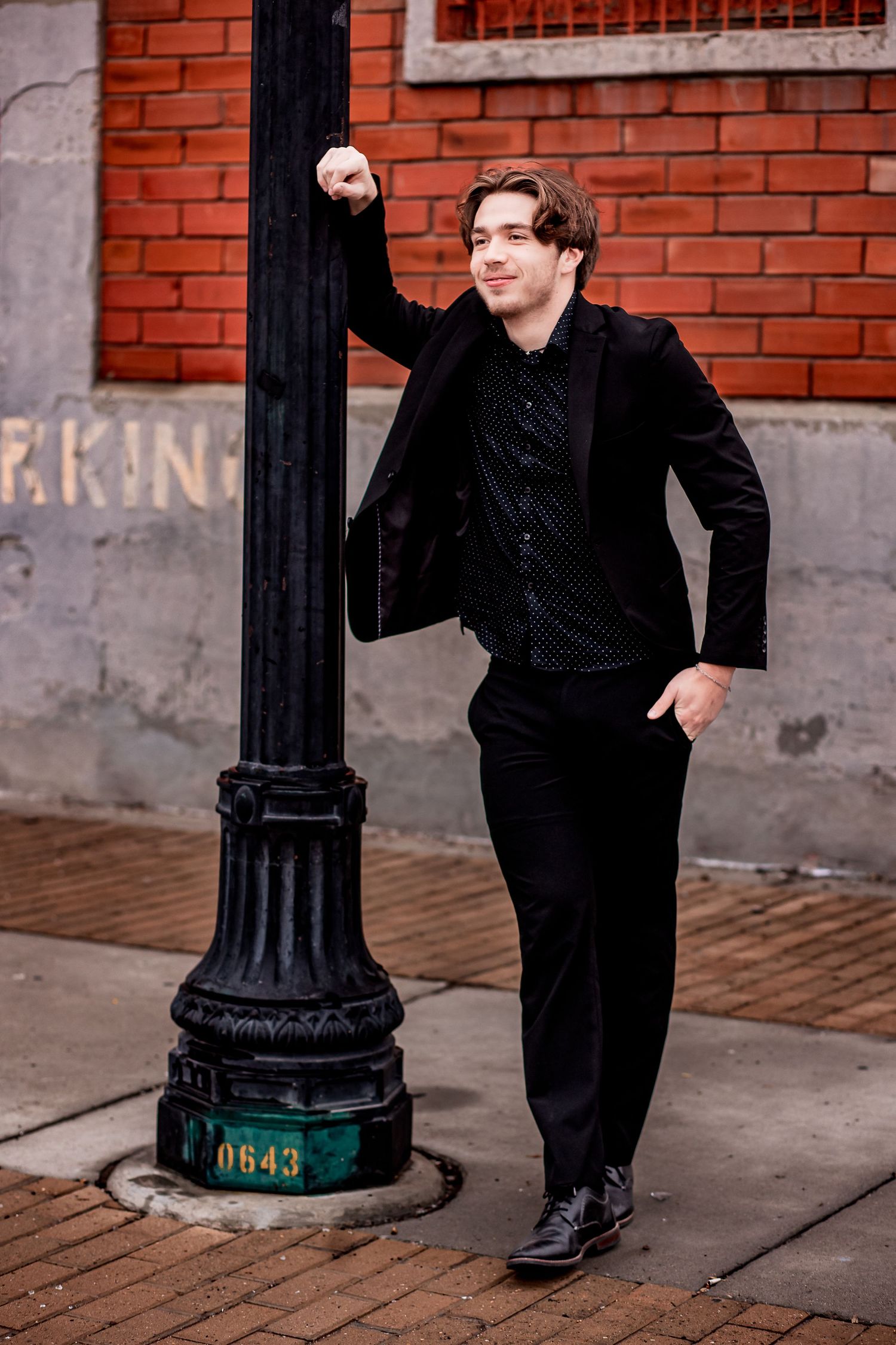 High school senior male wearing a sharp black suit leaning against a old fashioned light pole in a downtown urban environment.