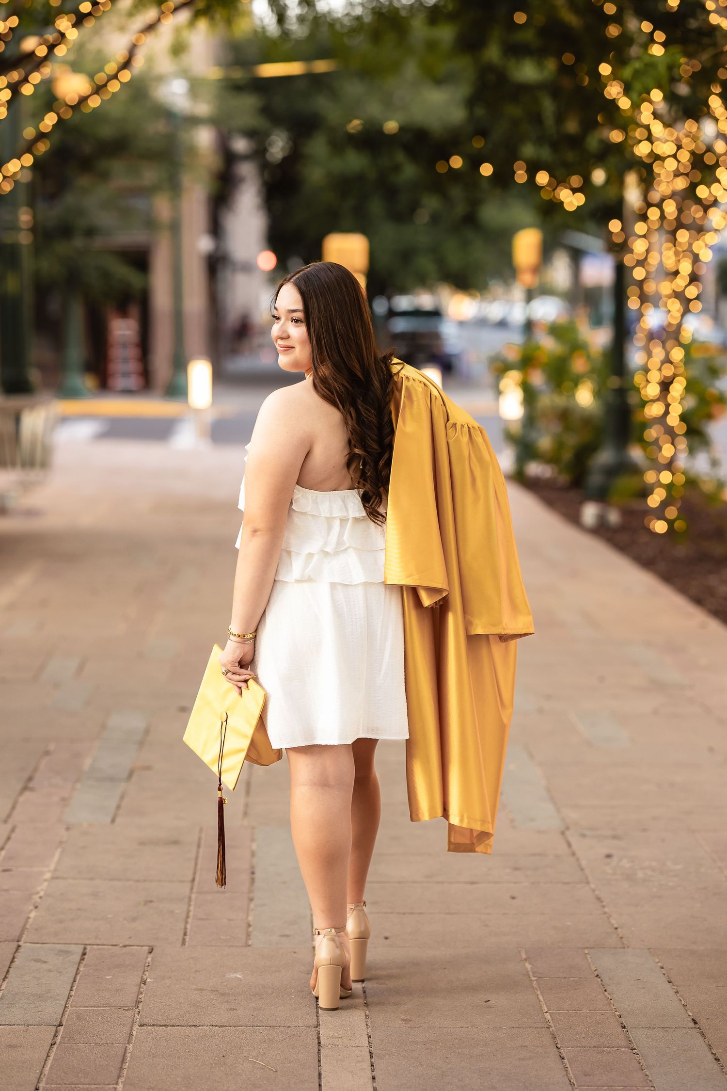 Female high school senior in a golden cap and gown celebrating graduation in downtown setting amongst sparkling lights.