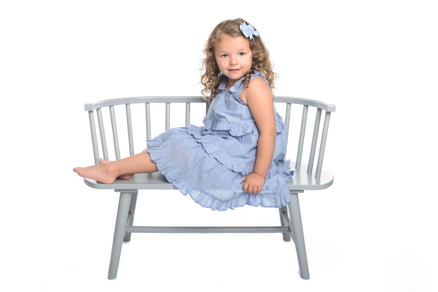 Child in blue ruffled dress posing on light gray bench in studio.
