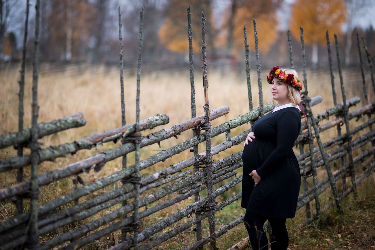 Pregnant woman in black dress and floral crown leaning against rustic wooden fence in autumn field.